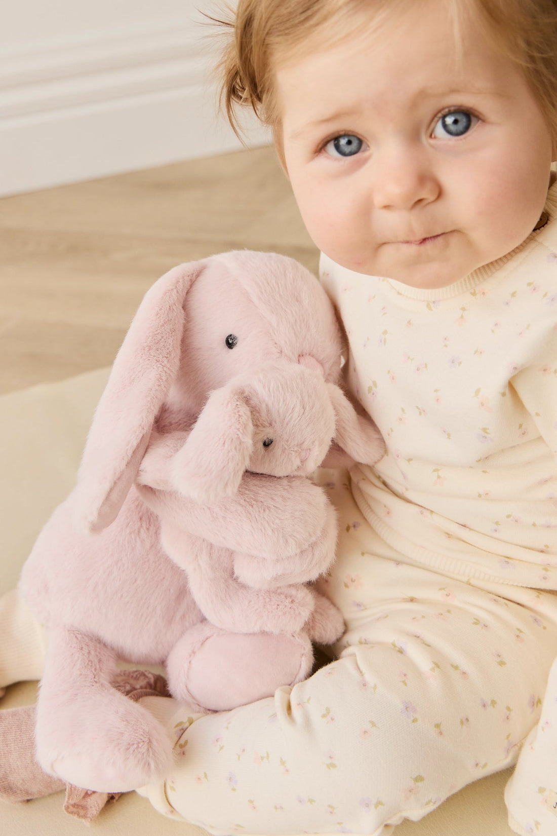 Baby holding pink plush bunny wearing floral patterned pajamas.