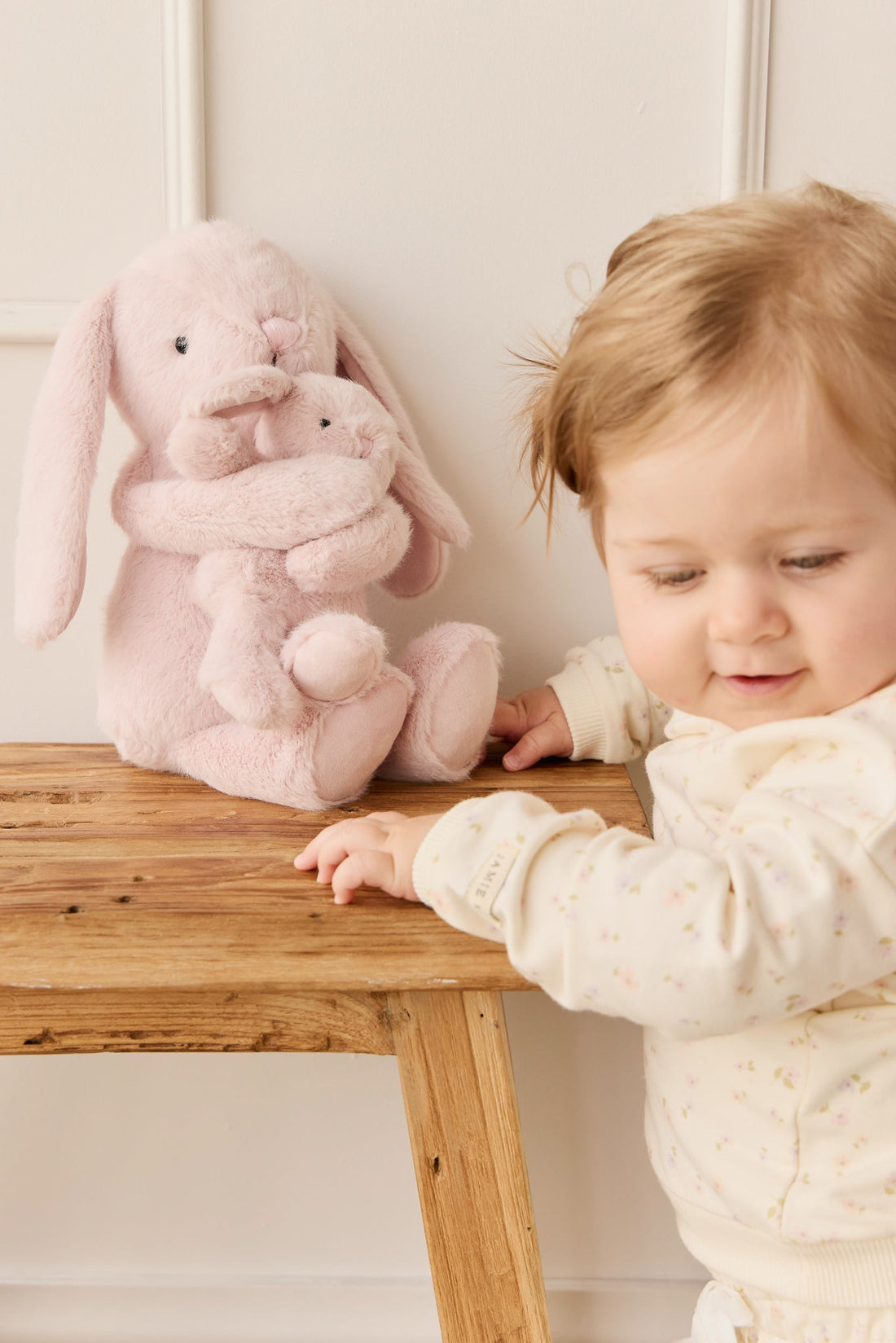 A baby reaching for a pink stuffed bunny on a wooden table.