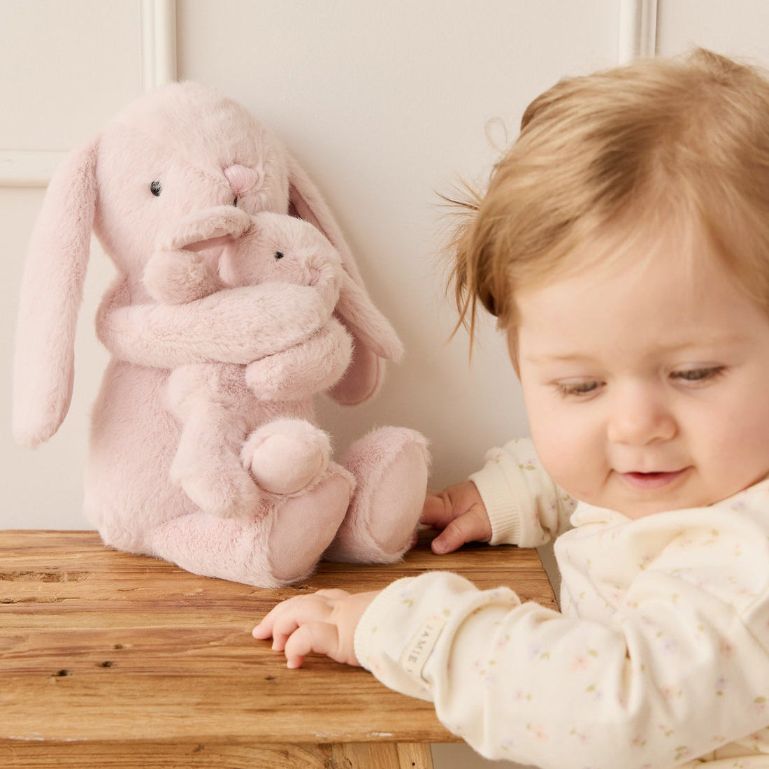 A baby reaching for a pink stuffed bunny on a wooden table.