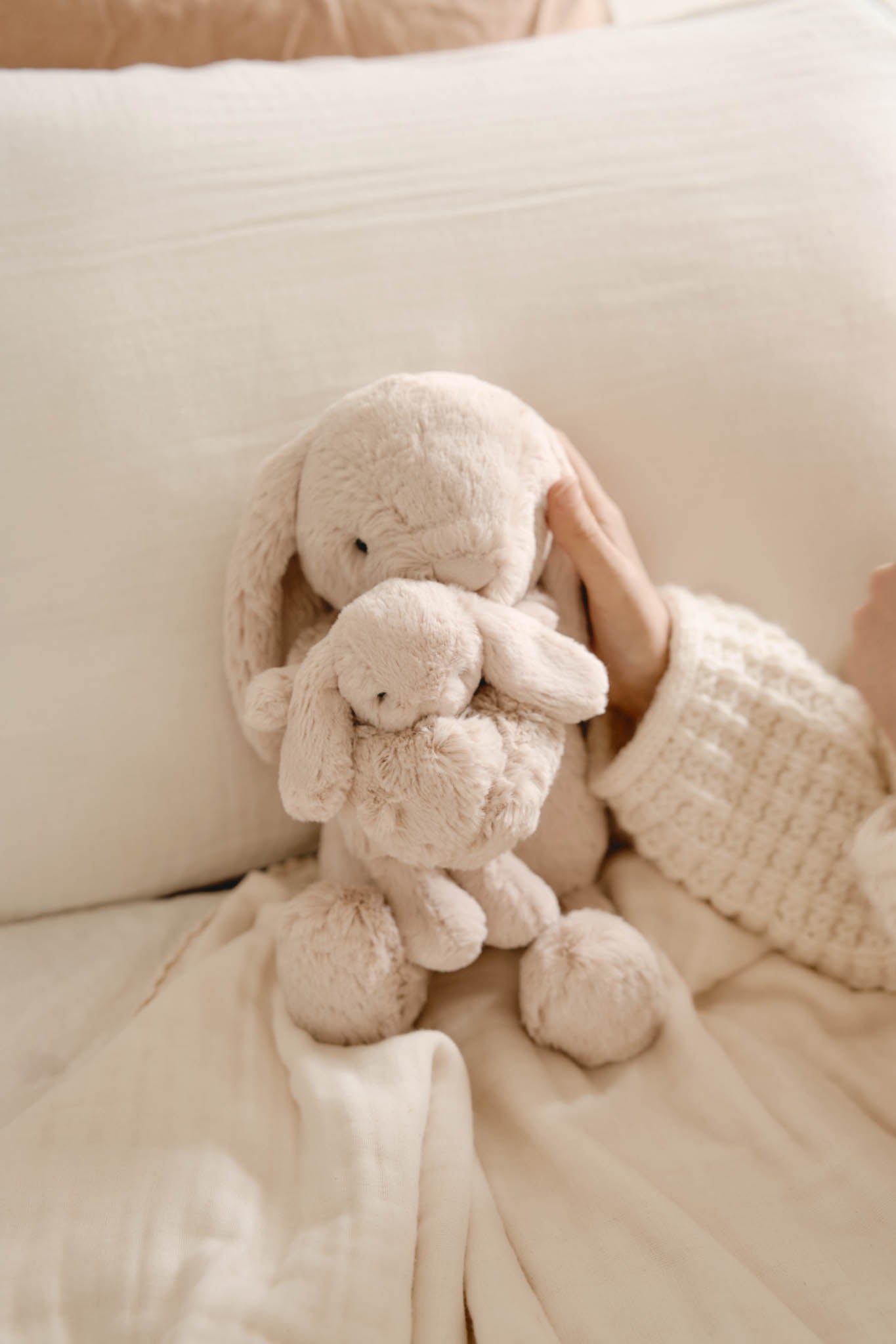 Two beige bunny plush toys stacked on a beige blanket next to a hand wearing a beige knit sweater.