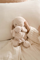Two beige bunny plush toys stacked on a beige blanket next to a hand wearing a beige knit sweater.