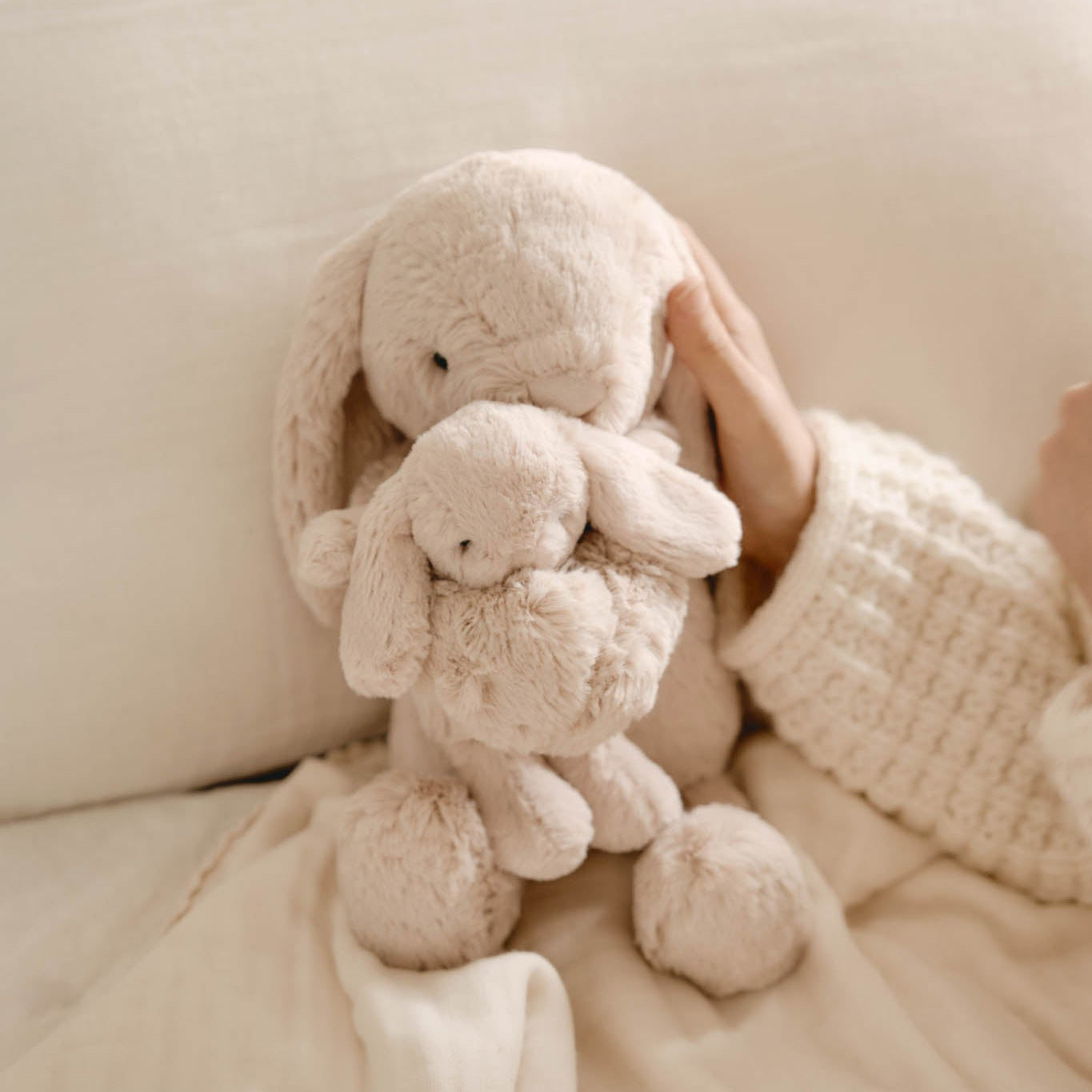 Two beige bunny plush toys stacked on a beige blanket next to a hand wearing a beige knit sweater.