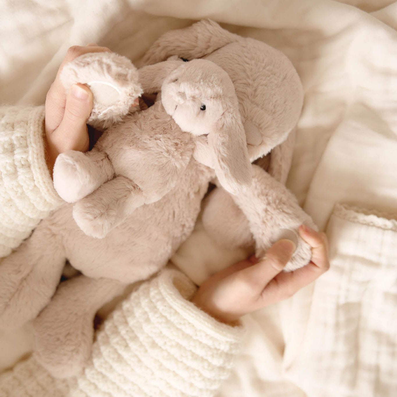Child's hands holding a beige stuffed rabbit toy on a white blanket, wearing a white cable-knit sweater.