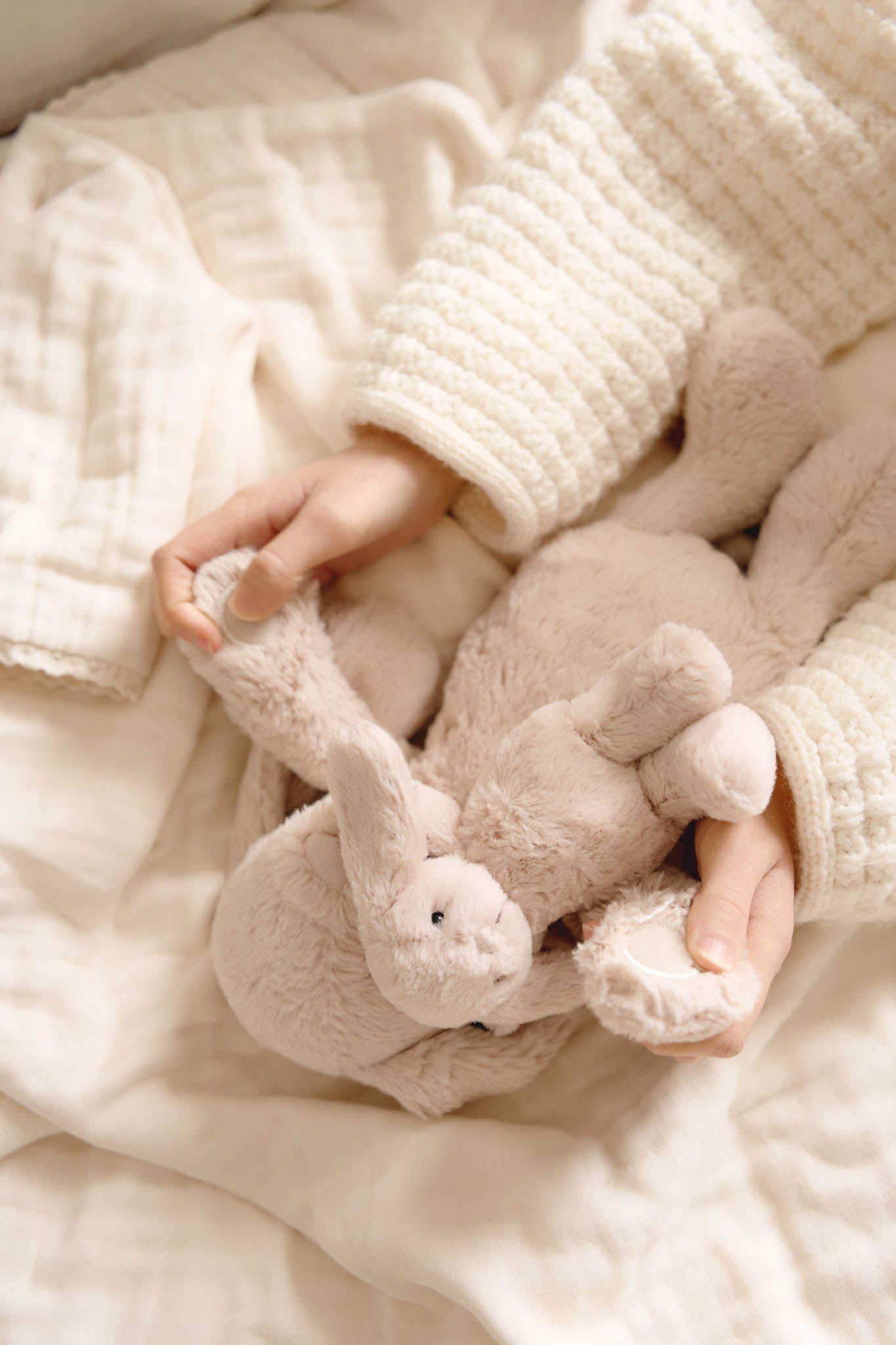 Child in a cream sweater holding a stuffed bunny in bed.