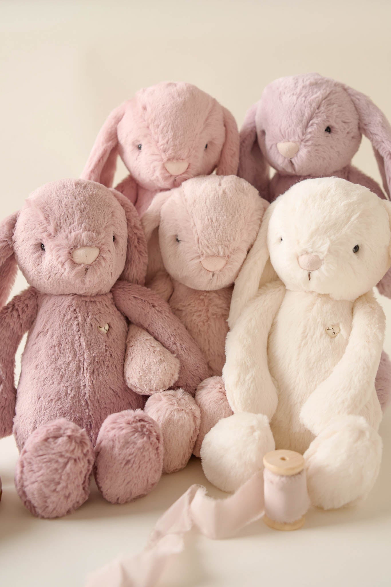 Five plush bunny toys in pastel pink, lavender, and white hues, grouped together with a spool of ribbon.