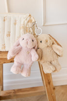 Two plush bunny keychain charms, pink and beige, hanging from a patterned bag on a wooden surface.