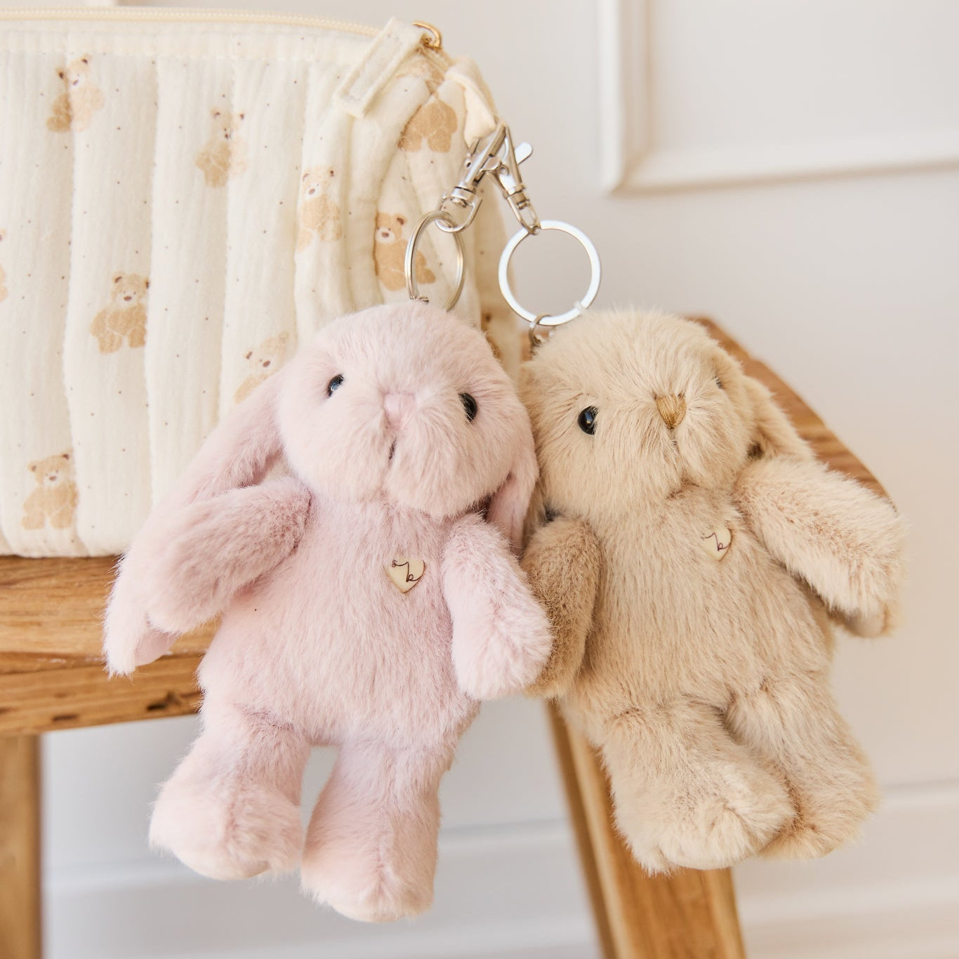Two plush bunny keychain charms, pink and beige, hanging from a patterned bag on a wooden surface.