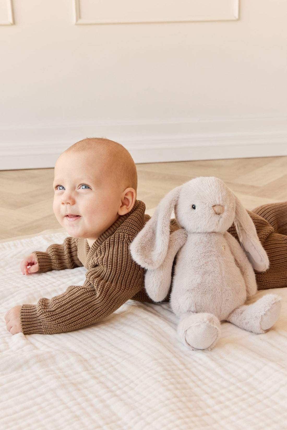 A baby in a brown knit outfit lying on a white blanket next to a plush rabbit.