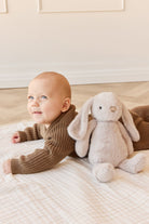 A baby in a brown knit outfit lying on a white blanket next to a plush rabbit.