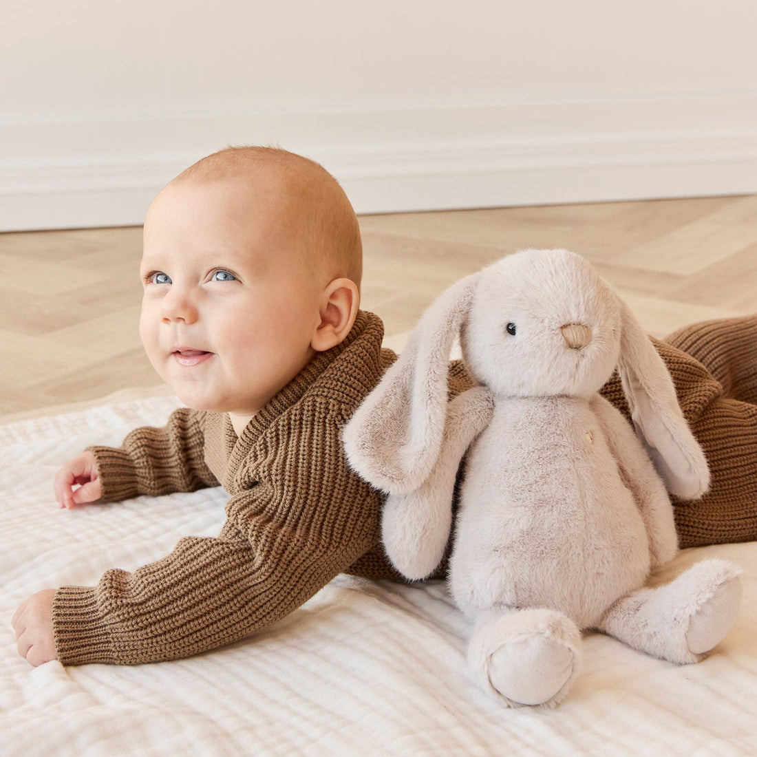 A baby in a brown knit outfit lying on a white blanket next to a plush rabbit.
