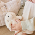A child holding a white plush bunny keychain in front of a ribbed beige bag. The child is wearing a white top and light green sweater.