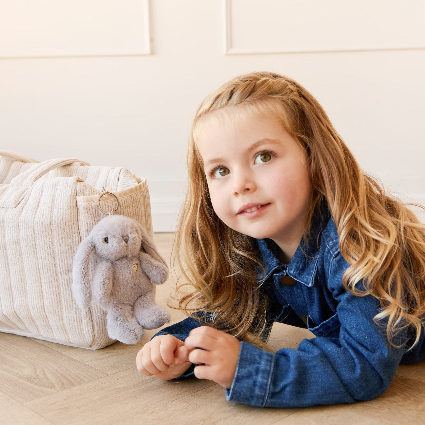 A young girl in a denim jacket poses with a plush bunny and a cream-colored corduroy bag.