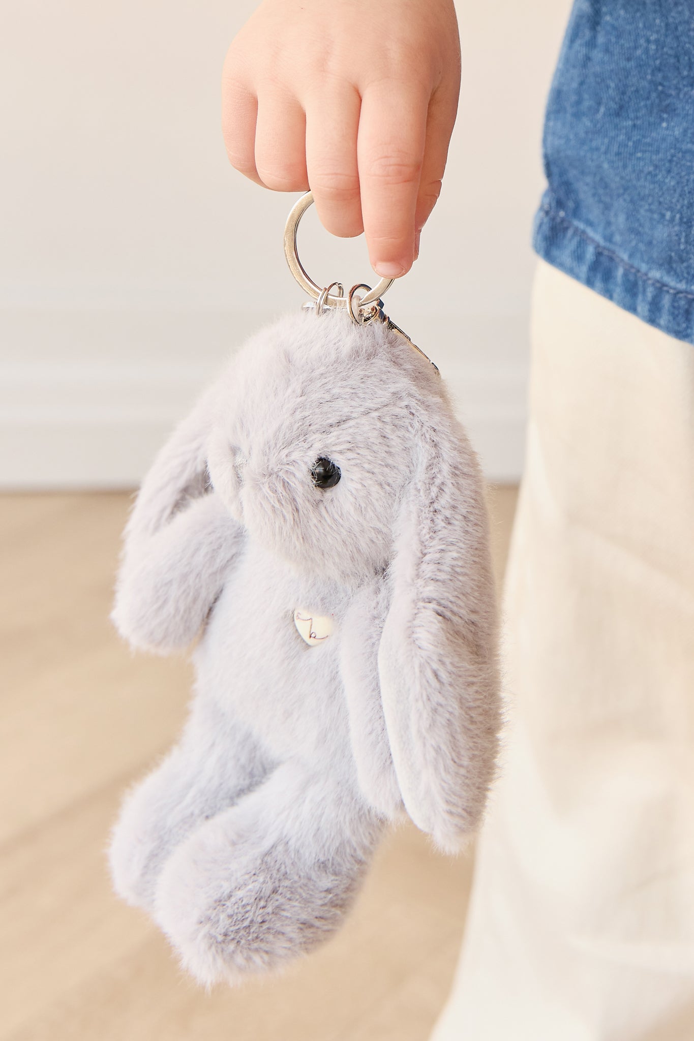 Child's hand holding a fluffy gray rabbit keychain, with a denim shirt and white pants visible.