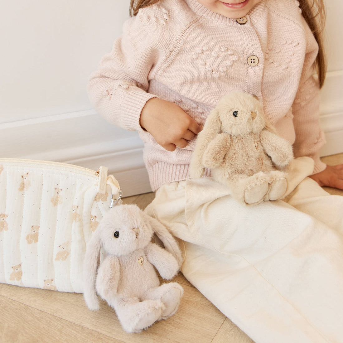 A child in a pink cardigan and cream-colored pants sits on the floor with two plush bunny toys and a cream-colored bag.