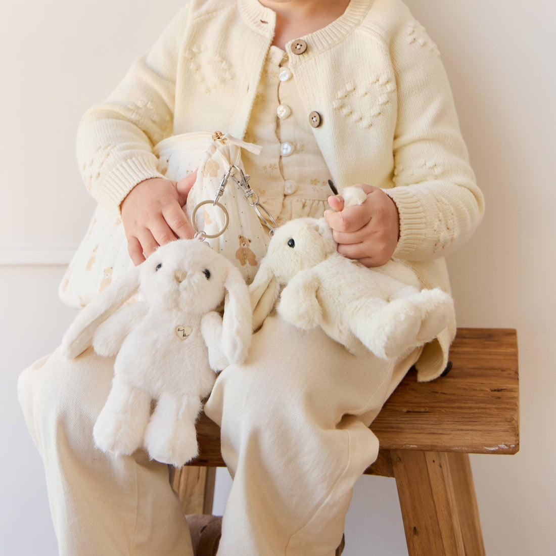 Child in a cream-colored knit cardigan with heart details and cream-colored pants, sitting on a wooden stool holding two stuffed bunnies.