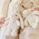 Two white bunny plush toys with heart-shaped buttons attached to metal clasps, held by a person wearing a cream-colored cardigan and pants.