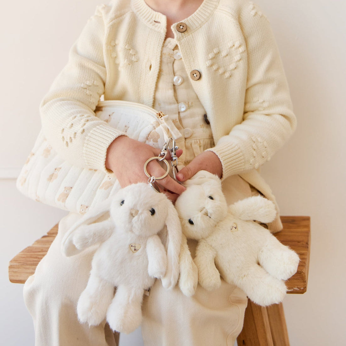 Young person in off-white outfit seated, holding plush bunny and bear keychains.
