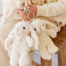 Two fuzzy white bunny keychains are held up by a person wearing a cream-colored cardigan.