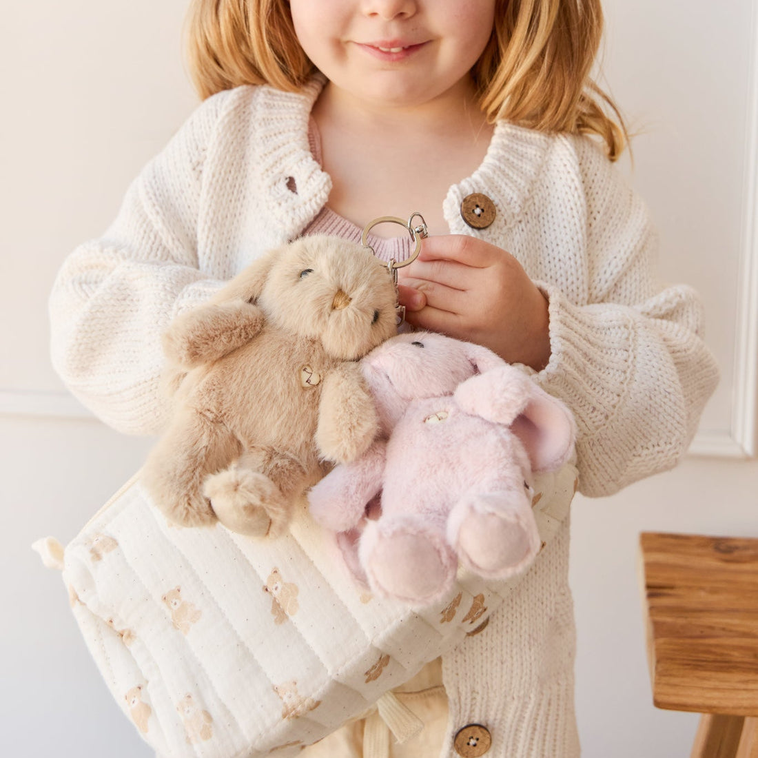 A young girl in a white cardigan holds a teddy bear patterned bag with two plush bunny keychains.
