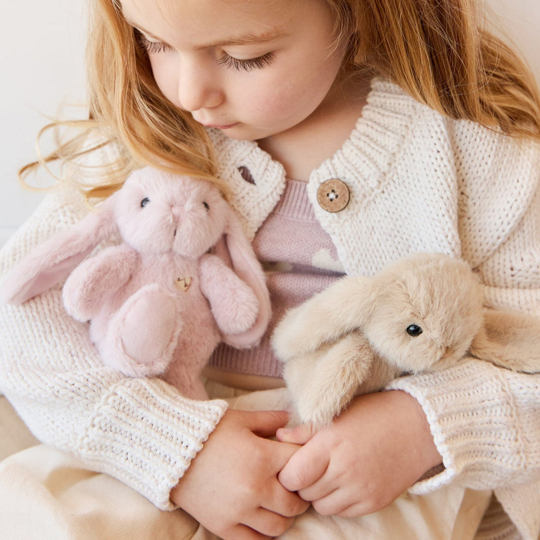 A young girl holds two plush bunnies while wearing a cream-colored knit cardigan.