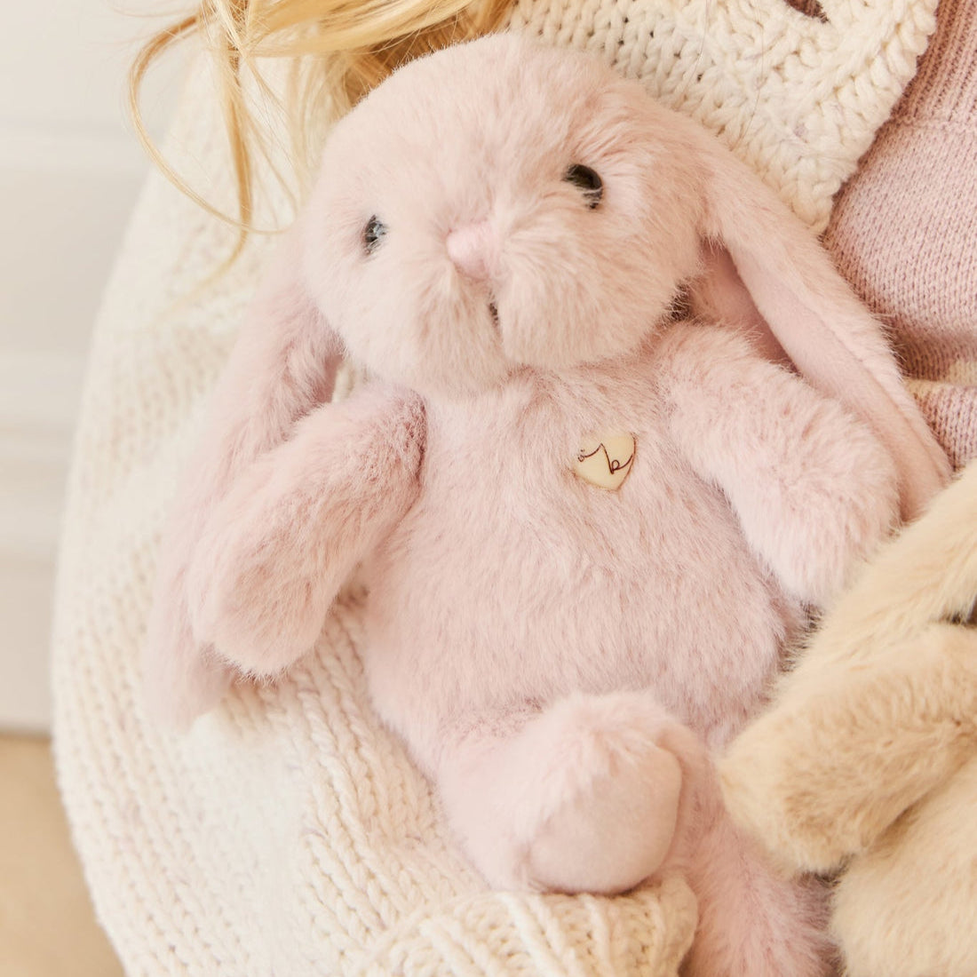 Pink plush bunny toy with wooden heart applique, nestled in white knit sweater.