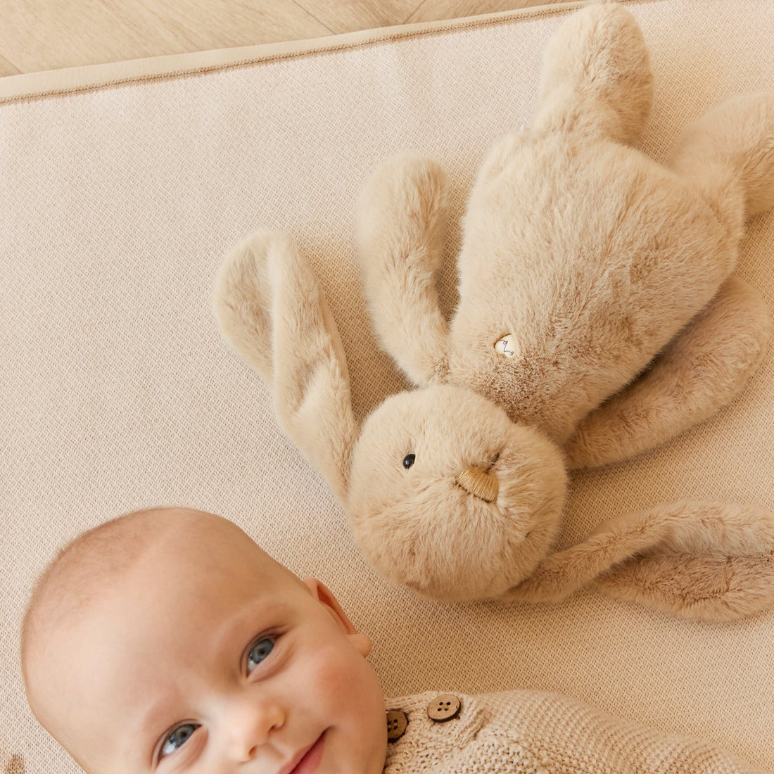 Baby in sweater and a beige plush bunny on a beige surface.