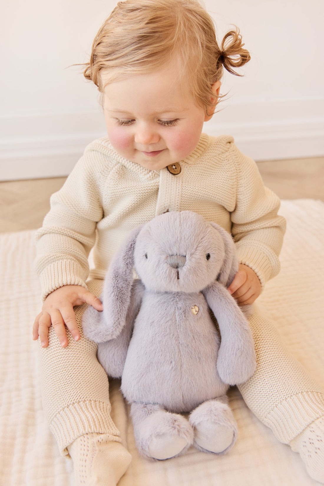 A child in a beige knit outfit sits with a gray plush rabbit.