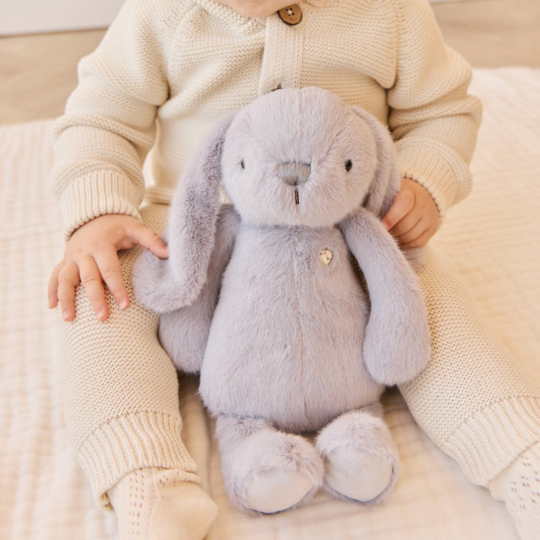 A child in a beige knit outfit sits with a gray plush rabbit.