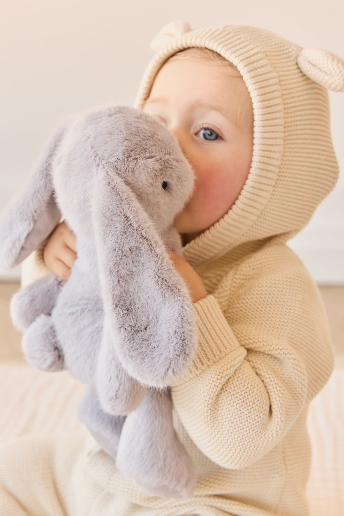 Toddler in a hooded sweater hugging a grey bunny stuffed animal.