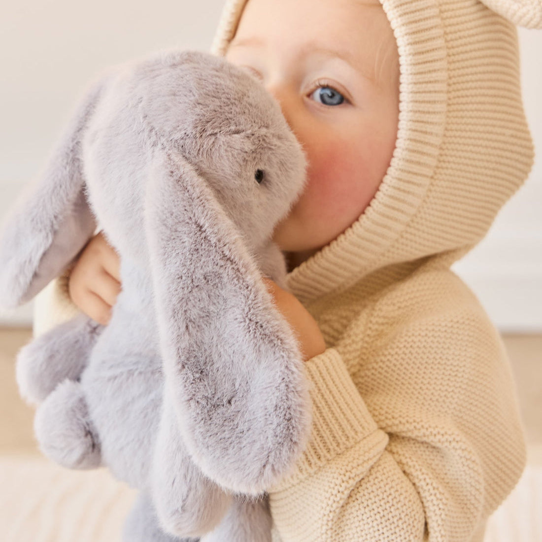 Toddler in a hooded sweater hugging a grey bunny stuffed animal.