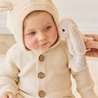 Baby wearing a knitted bunny-eared hooded cardigan with a plush bunny toy.