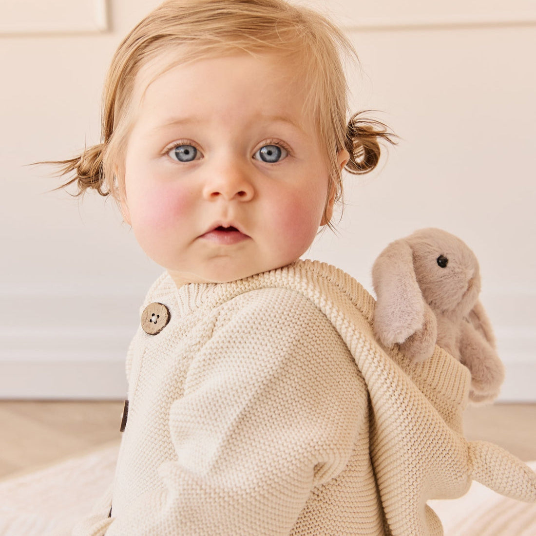 Baby with blue eyes, blonde pigtails, wearing a cream knit sweater, with a plush bunny.