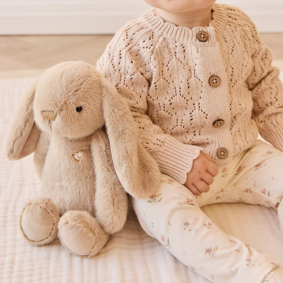 Child in open-knit cardigan and floral leggings, with plush bunny toy.