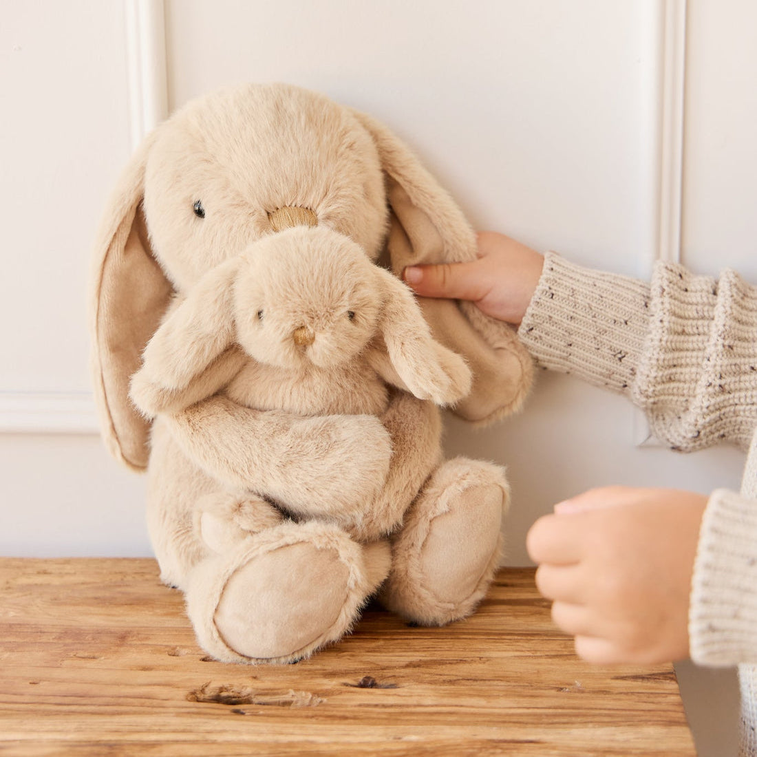 Two plush tan bunnies held by a child wearing a beige speckled sweater, sitting on a wooden surface.