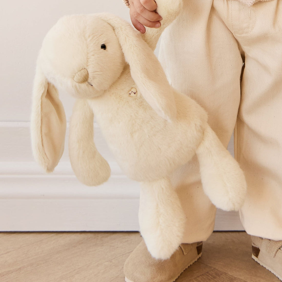 Child holding a plush bunny in a beige outfit and suede sandals.