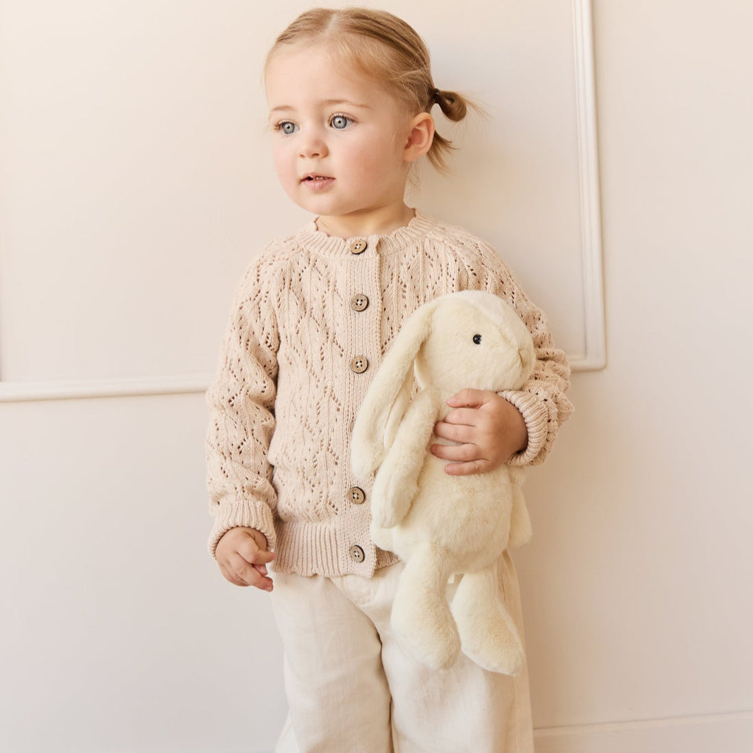A toddler wearing a beige knit cardigan and pants while holding a stuffed bunny.
