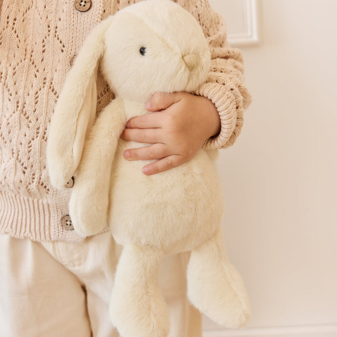 Child holding a cream-colored plush bunny, wearing a beige knit cardigan and pants.