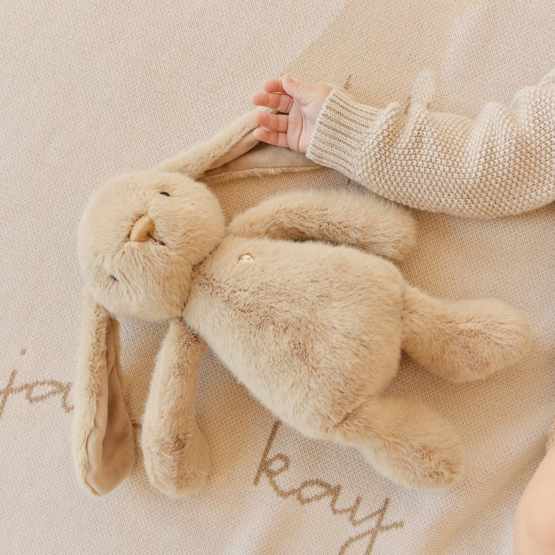 Tan plush bunny with a baby's hand on a blanket.