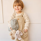 A child in beige overalls and a knitted sweater holds a quilted backpack with bunny keychains.
