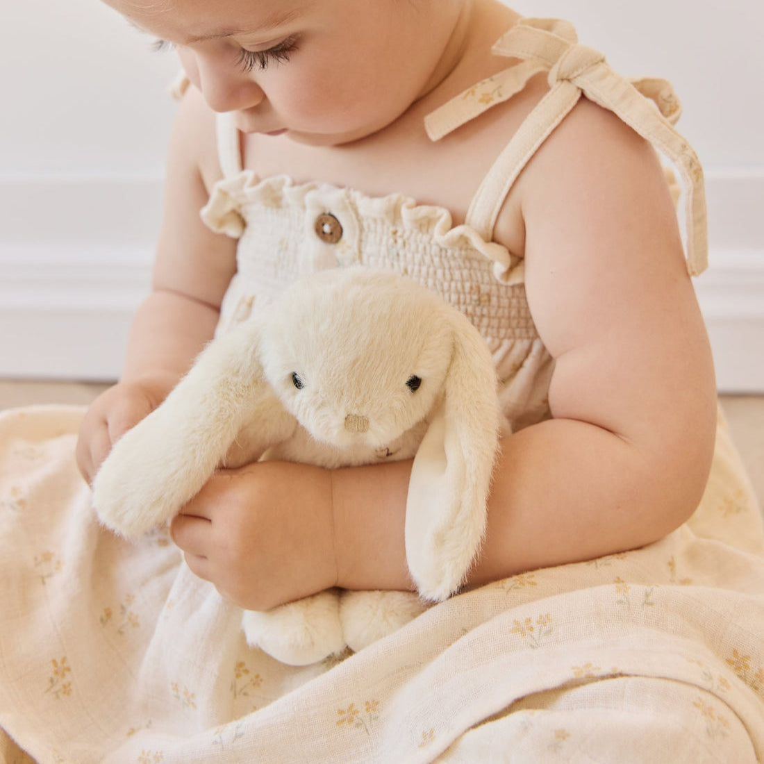 A toddler holding a stuffed bunny in a beige dress.
