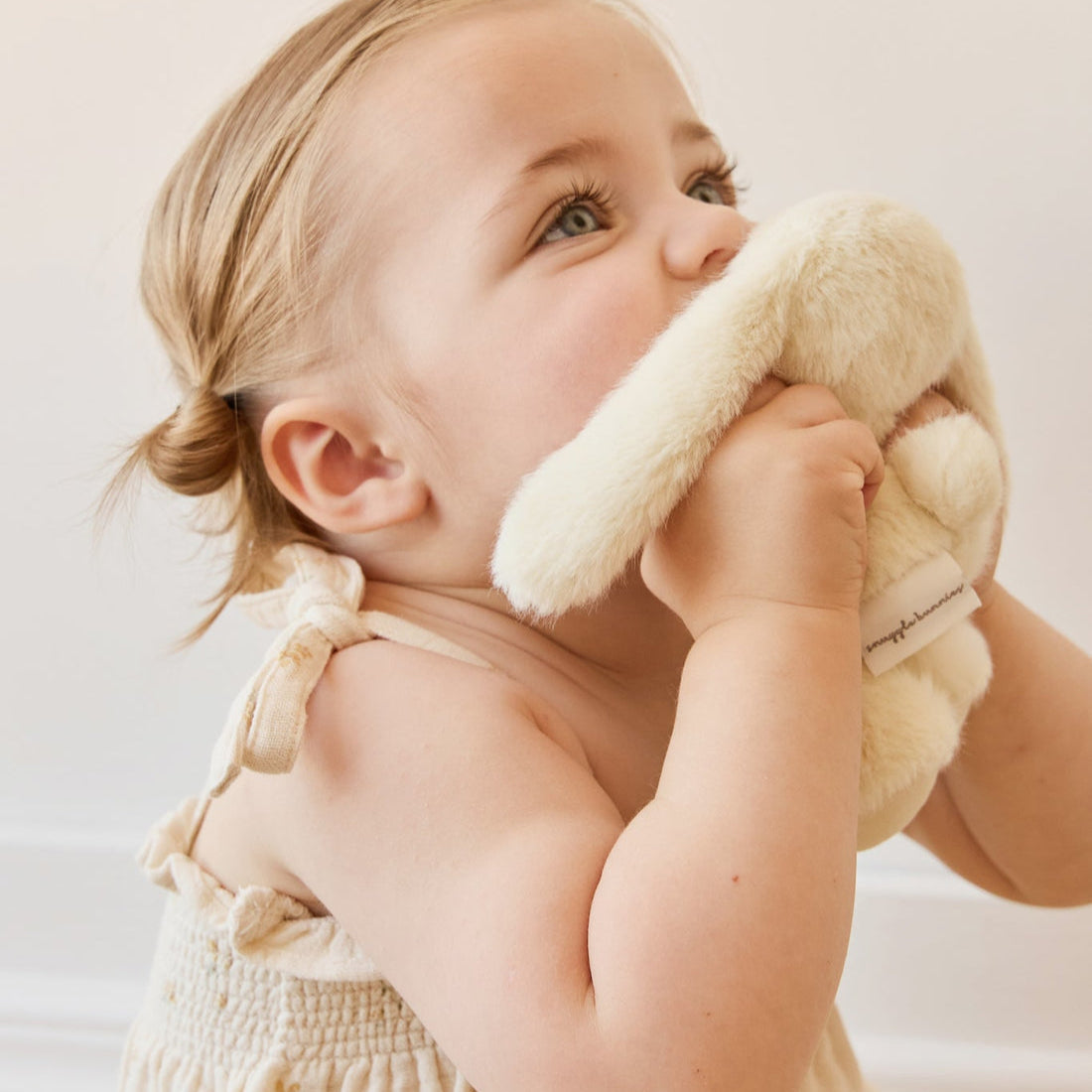 A baby in a beige dress holding a white plush bunny.