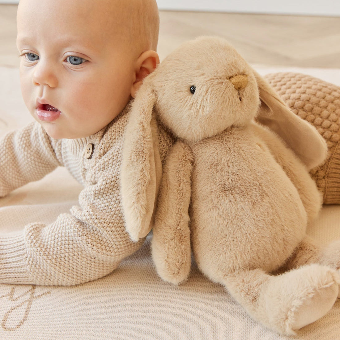 Baby wearing beige knit sweater and laying next to stuffed rabbit toy.