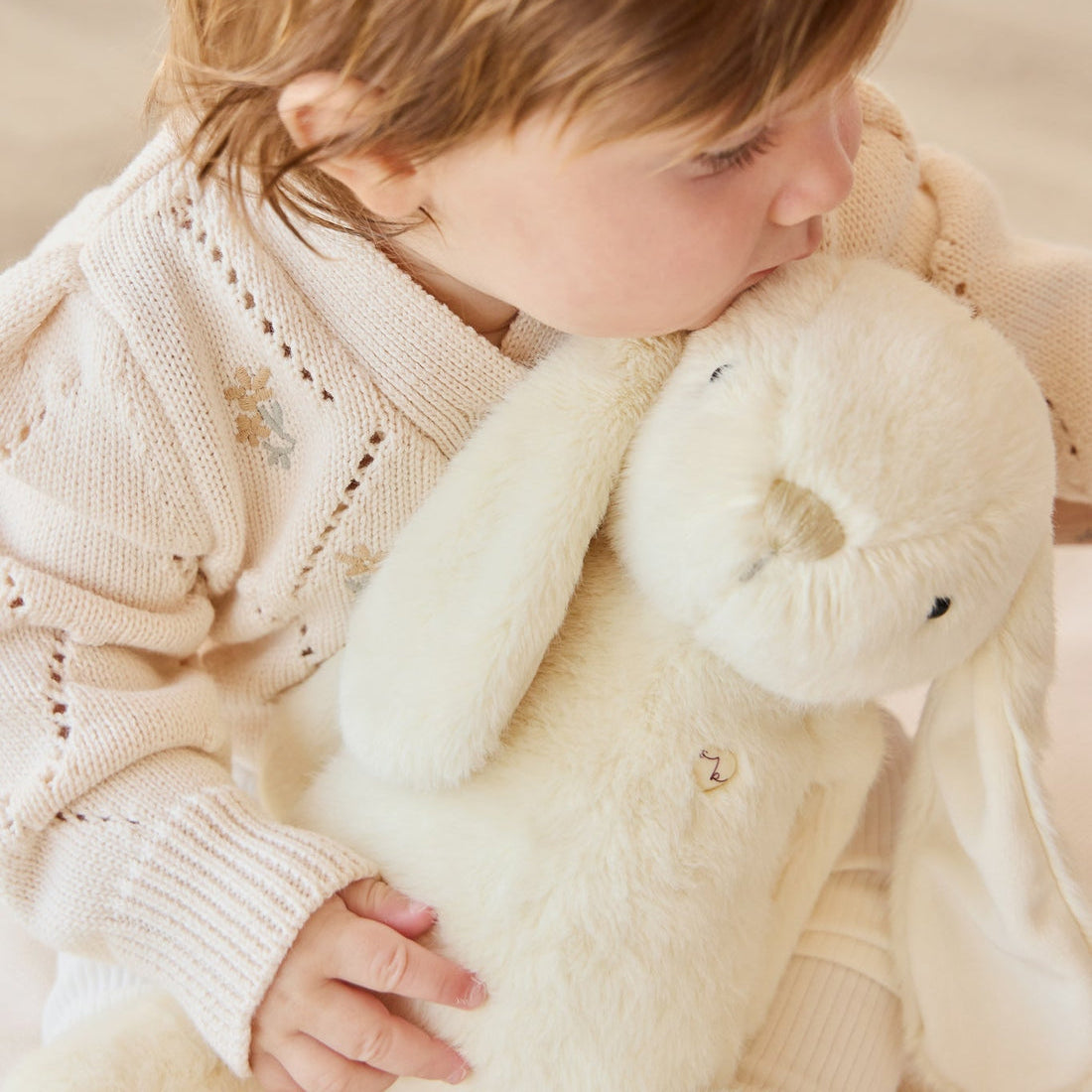 Child hugging stuffed white rabbit, wearing embroidered knit sweater.