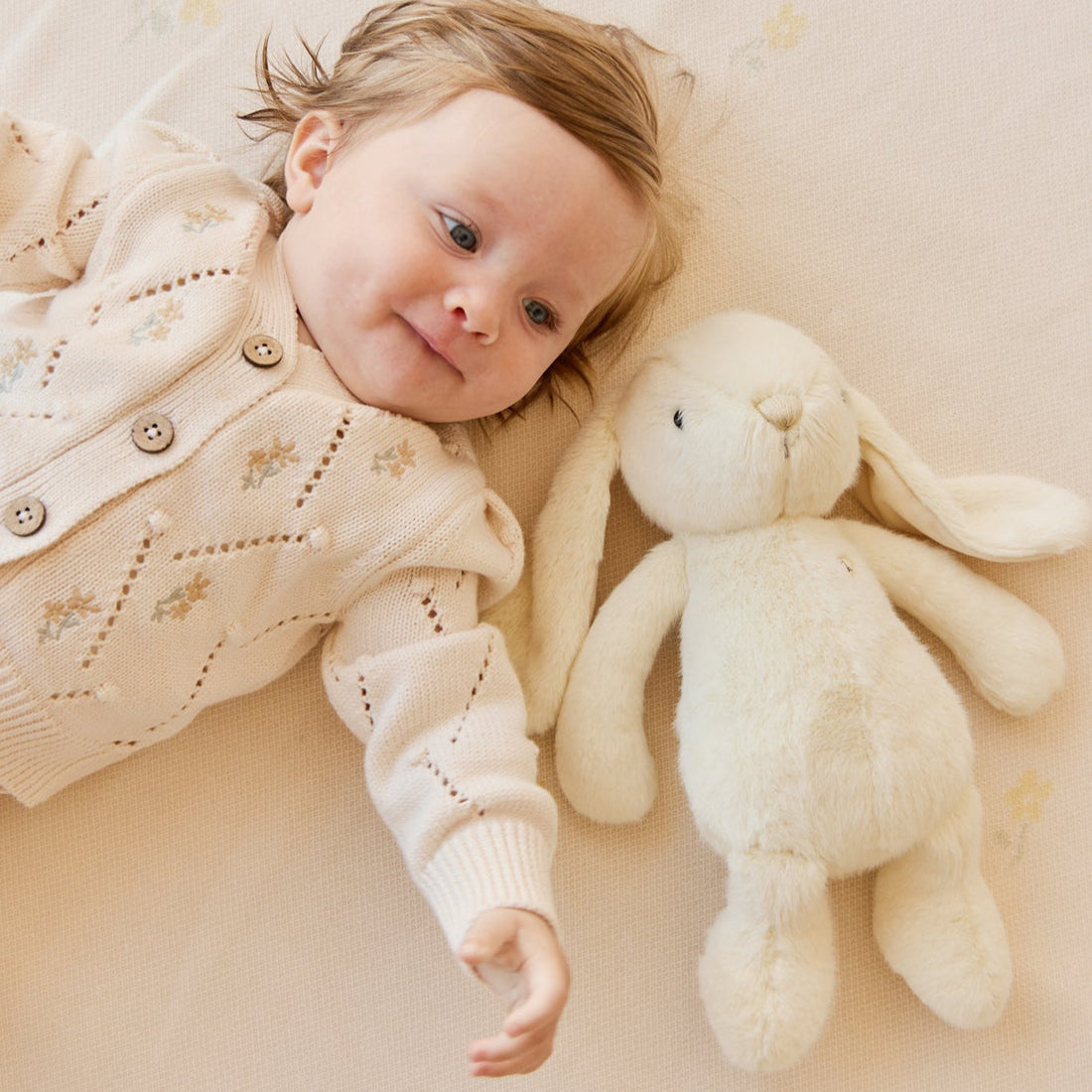 Baby in knit cardigan lying next to stuffed bunny.
