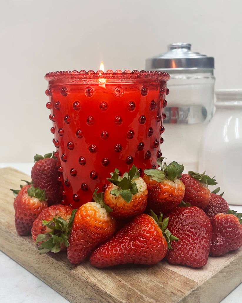 Red candle with a textured surface surrounded by strawberries on a wooden board.
