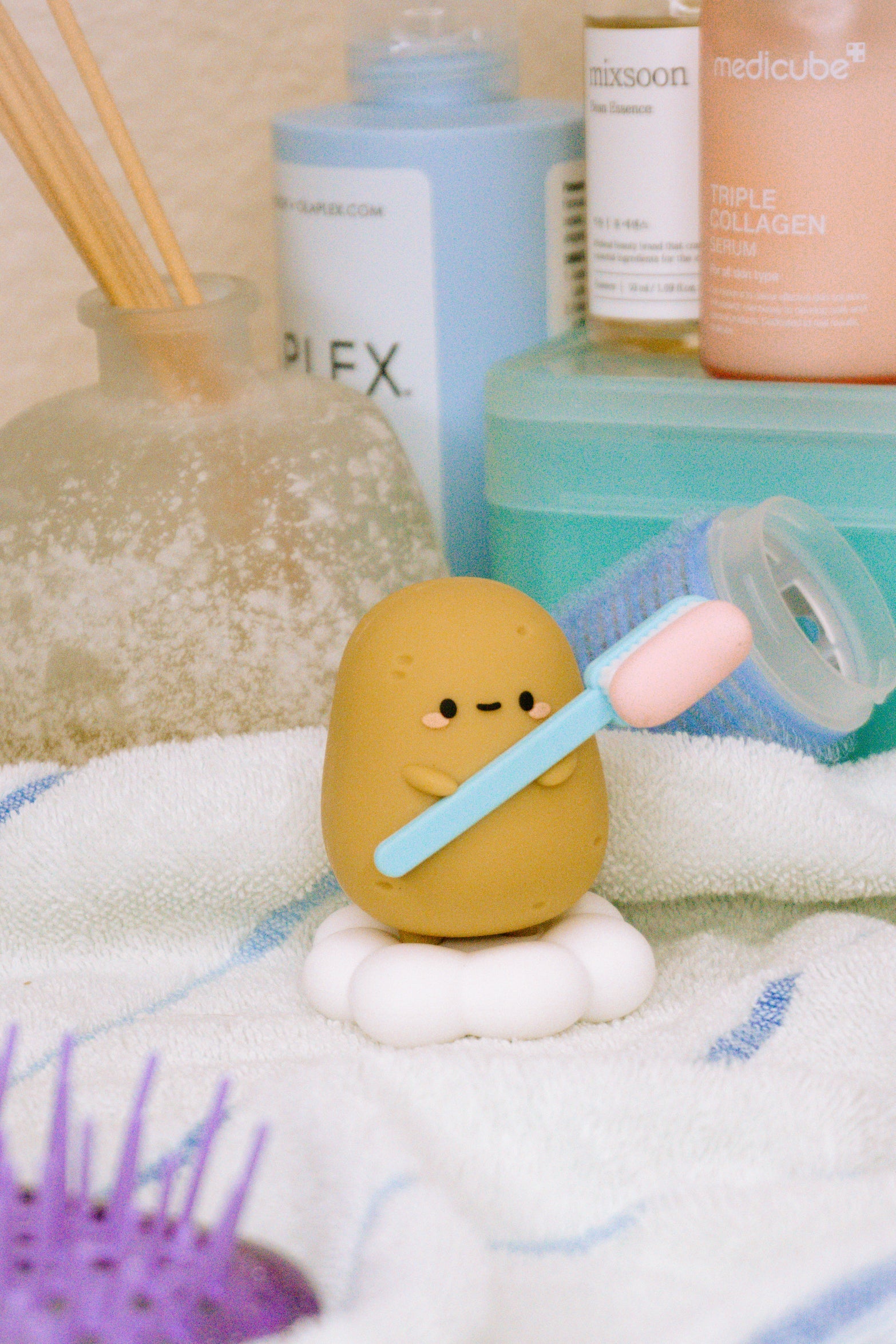 Potato-shaped figurine holding blue and pink toothbrush, sitting on white cloud. Skincare products and diffuser in background. White and blue towel below. Purple hairbrush visible at bottom.