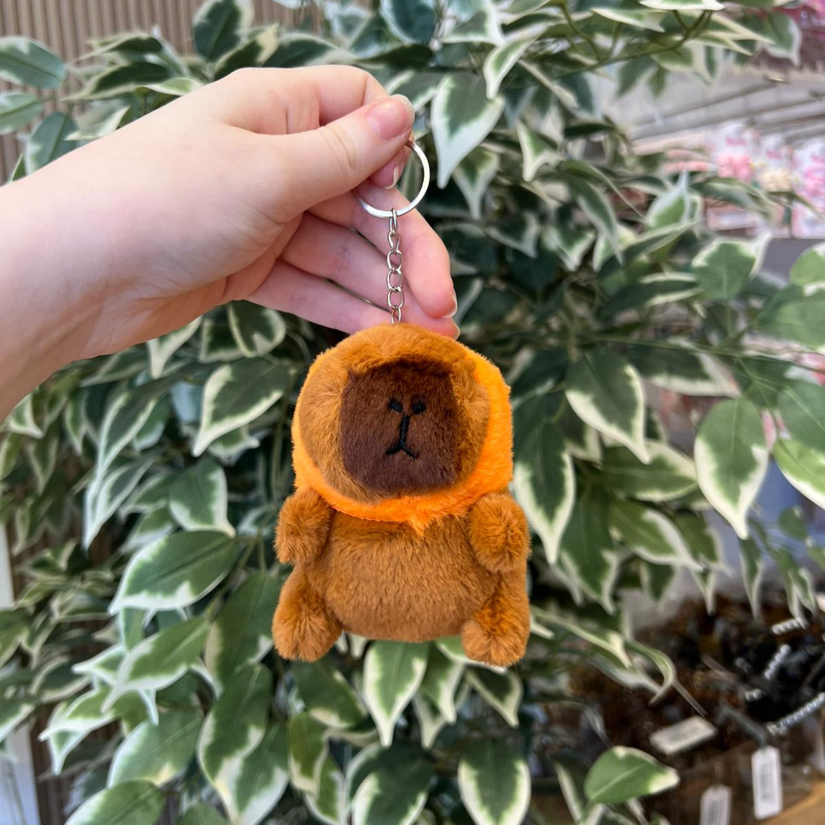 A hand holds a brown plush bear keychain with an orange collar against a leafy background.