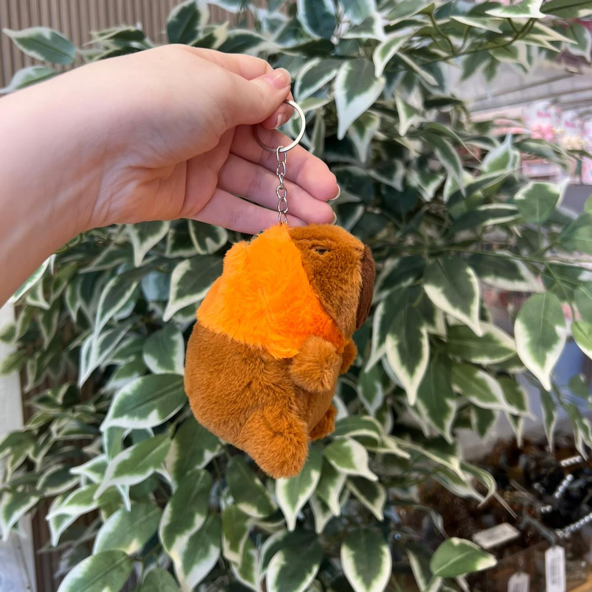 Hand holding a brown stuffed animal keychain with orange accents, against a backdrop of green and white variegated leaves.