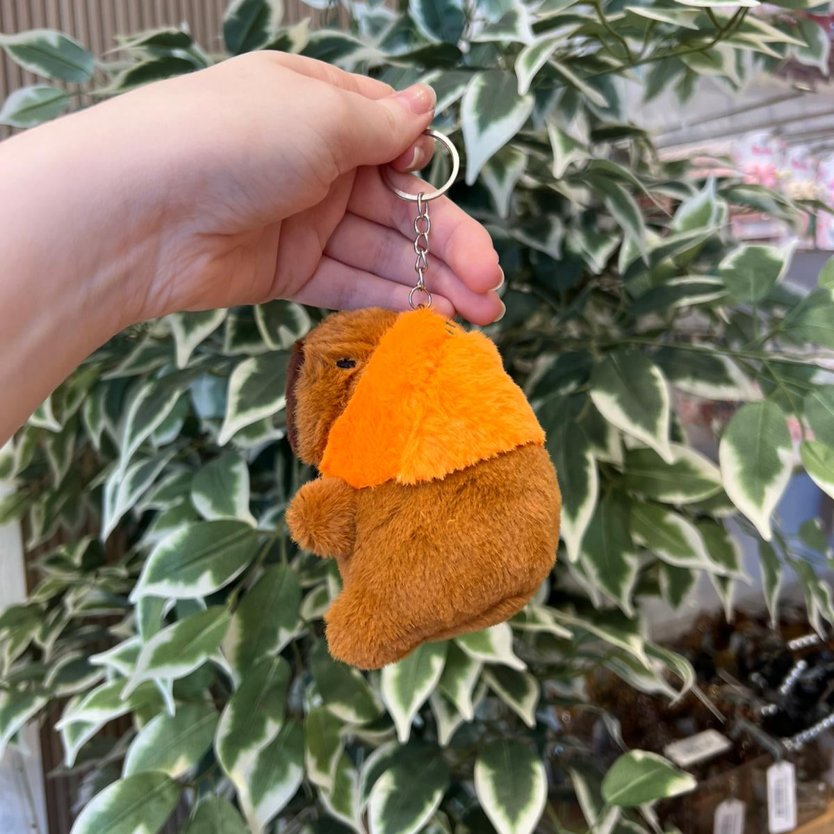 A hand holds a brown plush dachshund keychain with bright orange floppy ears against a background of green and white variegated leaves.
