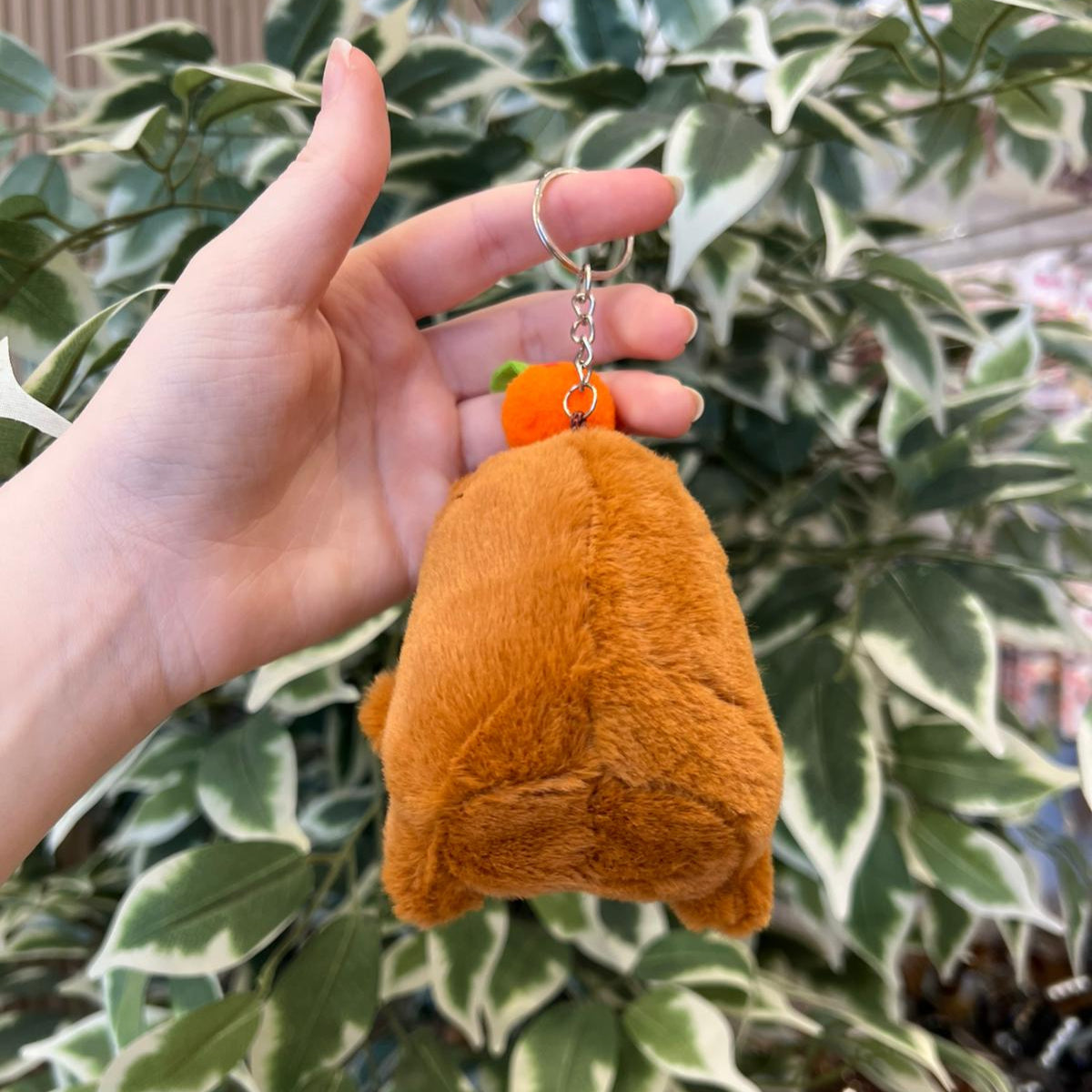 A hand holds a brown, plush bear keychain with an orange ball on top against a leafy green background.